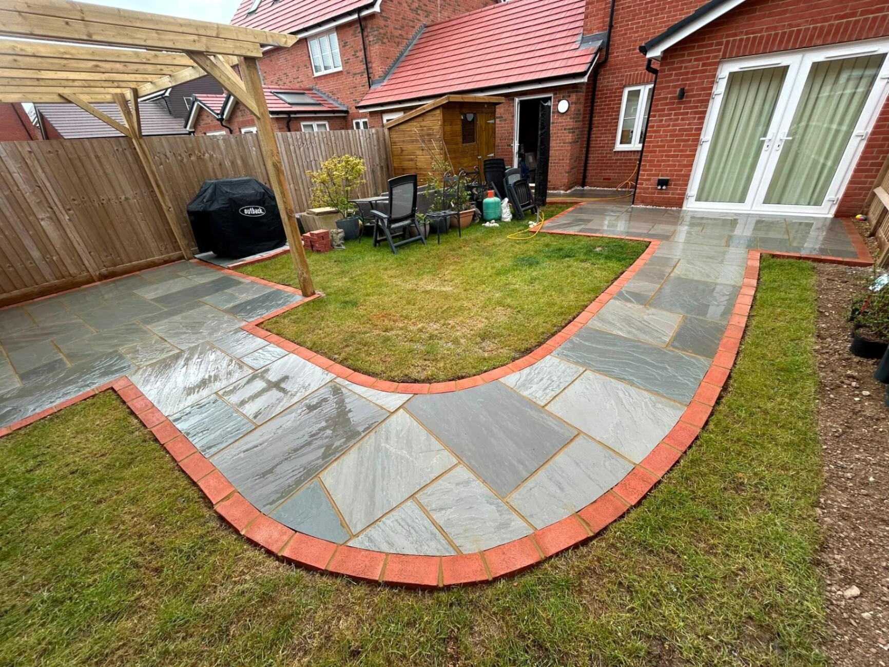 Indian sandstone patio with red-brick edging curving around a manicured rear lawn — a recent Heavens Property Solutions patio installation in West Sussex.